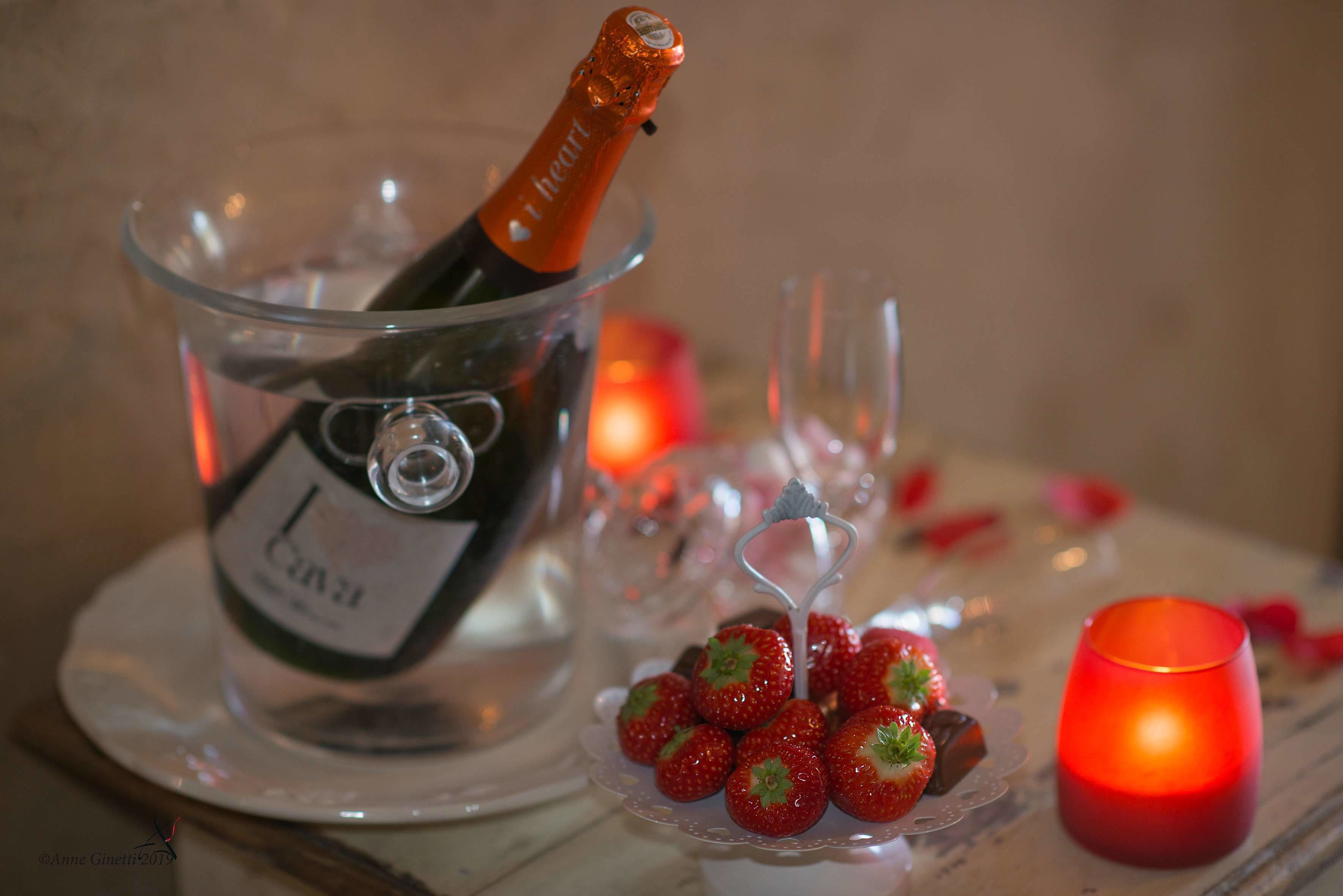 A bottle of champagne in a glass bowl, strawberries on a small plate, and a lit candle on a table.