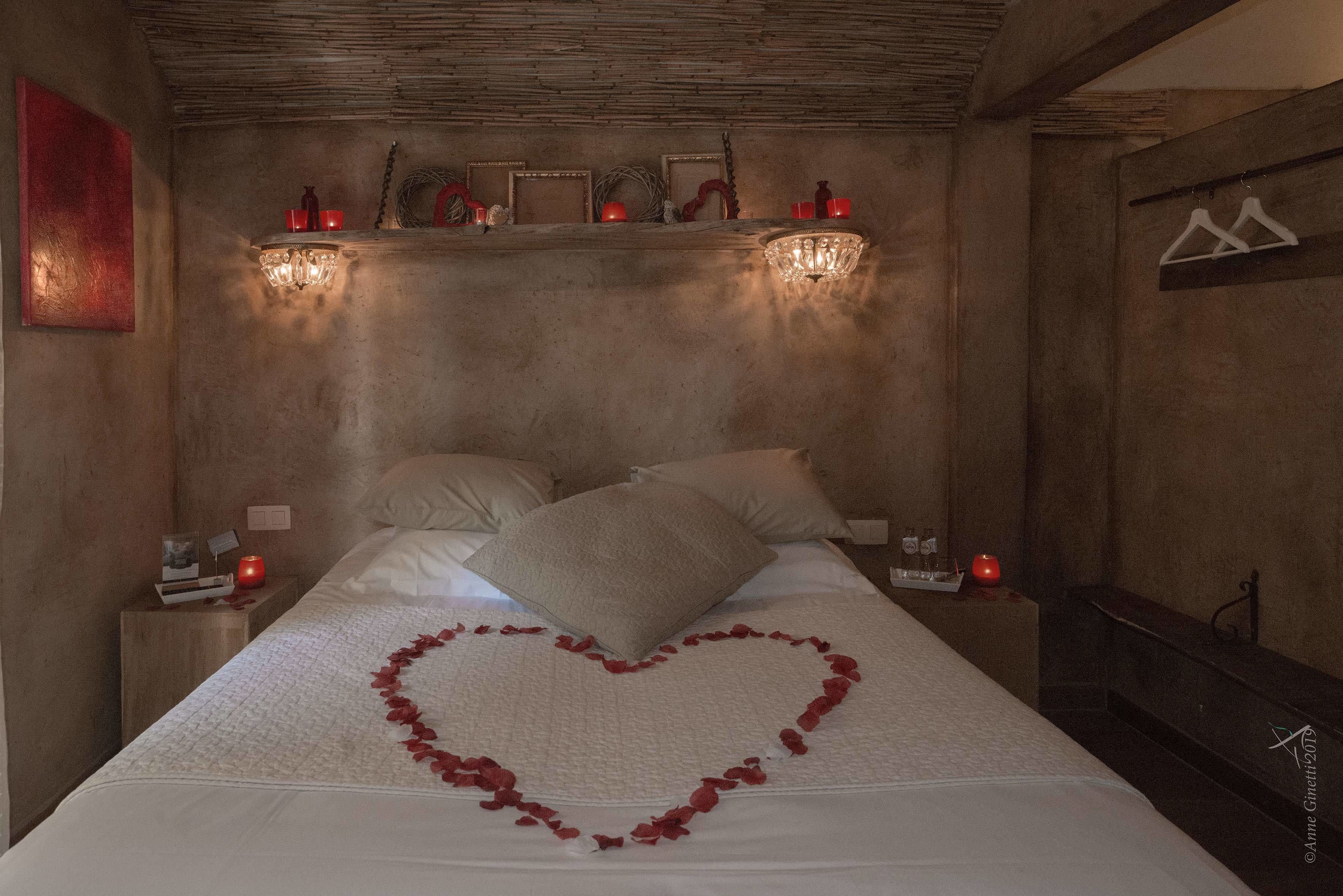 A bedroom with a bed, pillows, a heart-shaped decoration on the bed, wall sconces, a shelf with decorative items, and a coat rack.