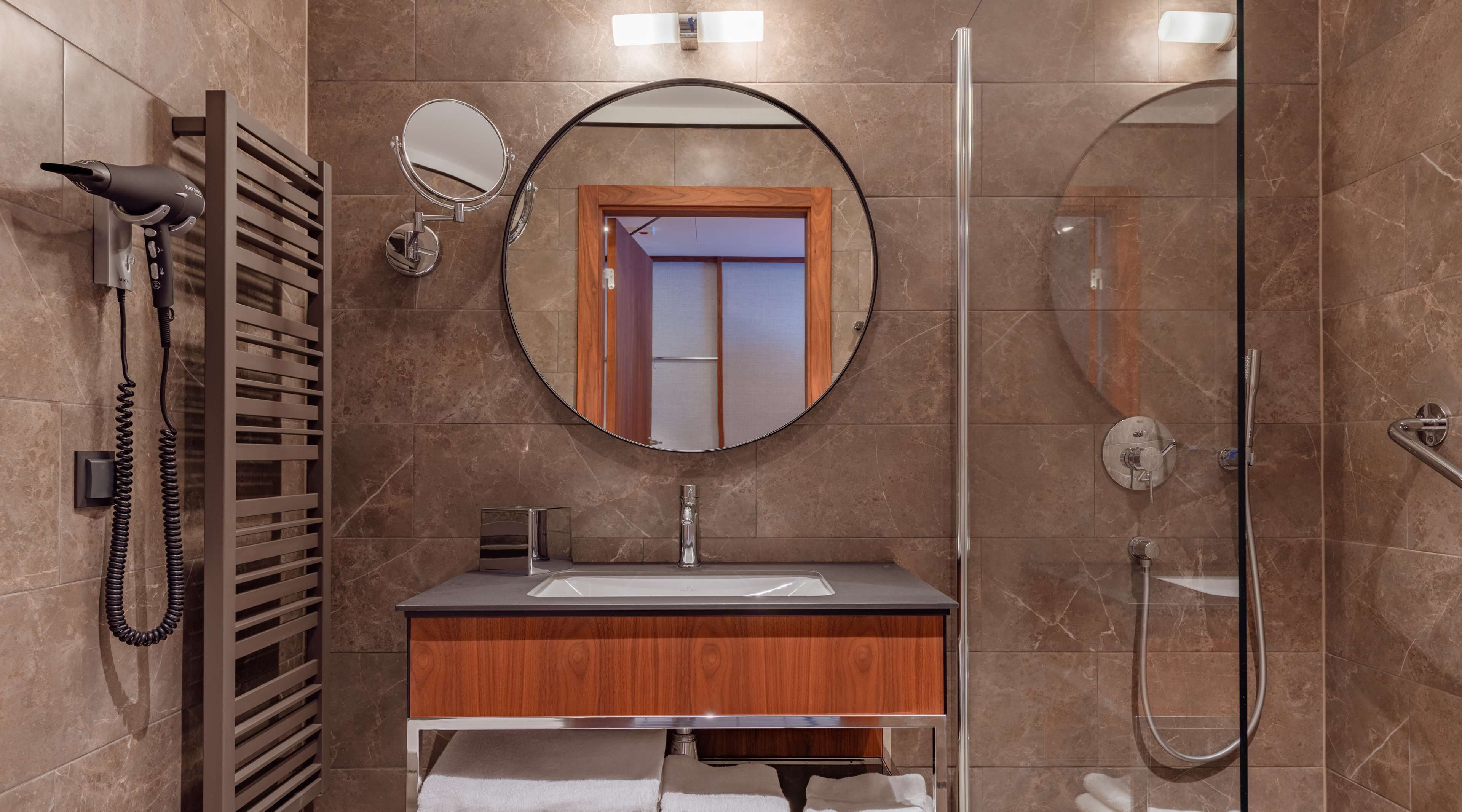 A modern bathroom with a wooden vanity, a round mirror, a hairdryer, and a glass-enclosed shower.