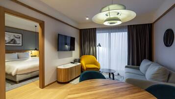 Ein Hotelzimmer mit einem Bett, einem Sofa, einem runden Holztisch, einem gelben Sessel, einem Fernseher und einem großen Fenster mit Vorhängen.