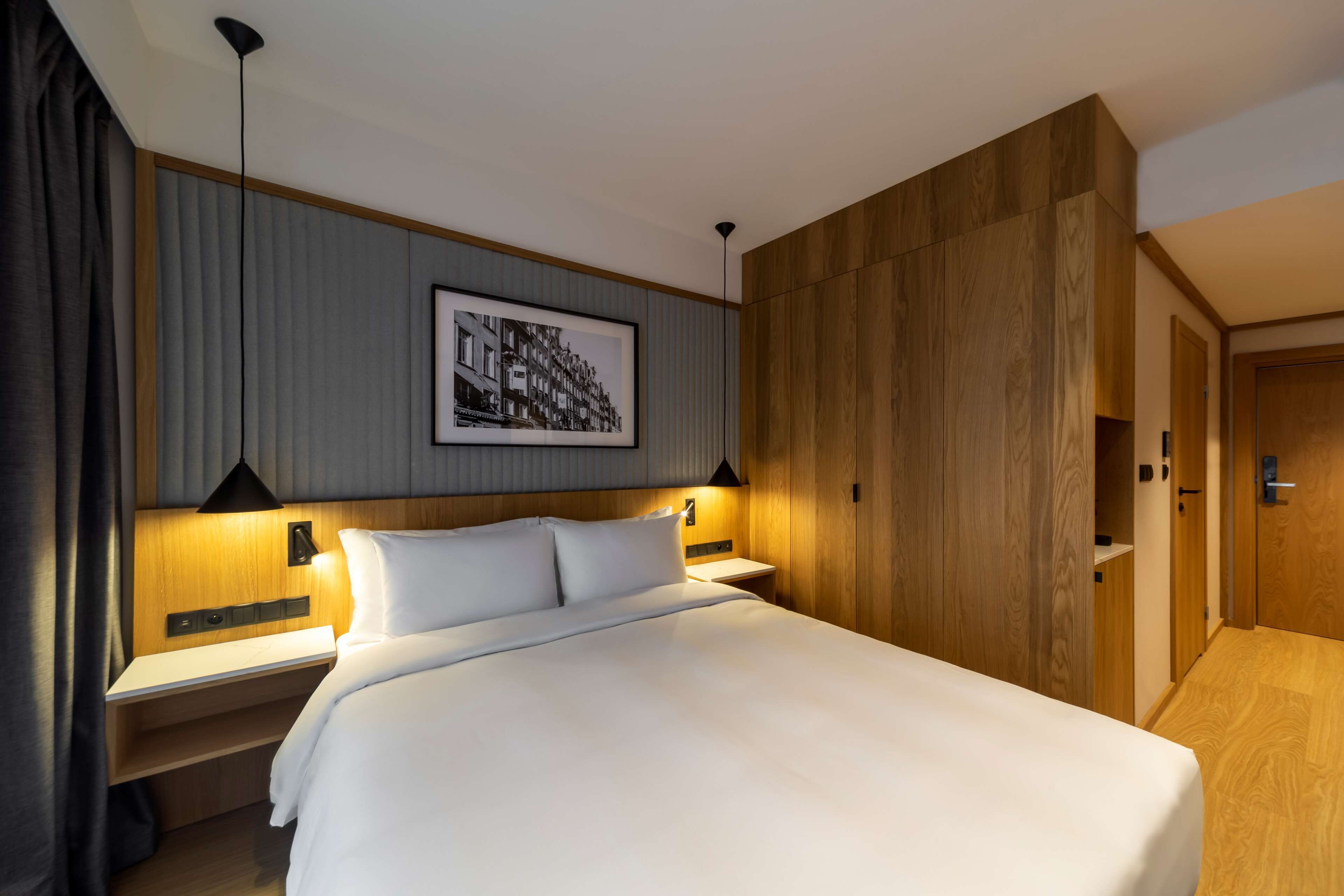 A modern hotel room with a large bed, wooden paneling, a framed cityscape picture, and a nightstand with a lamp.