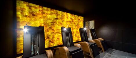 A row of massage chairs with wooden armrests and black seats, positioned against a wall with a textured, warm-toned mosaic design.