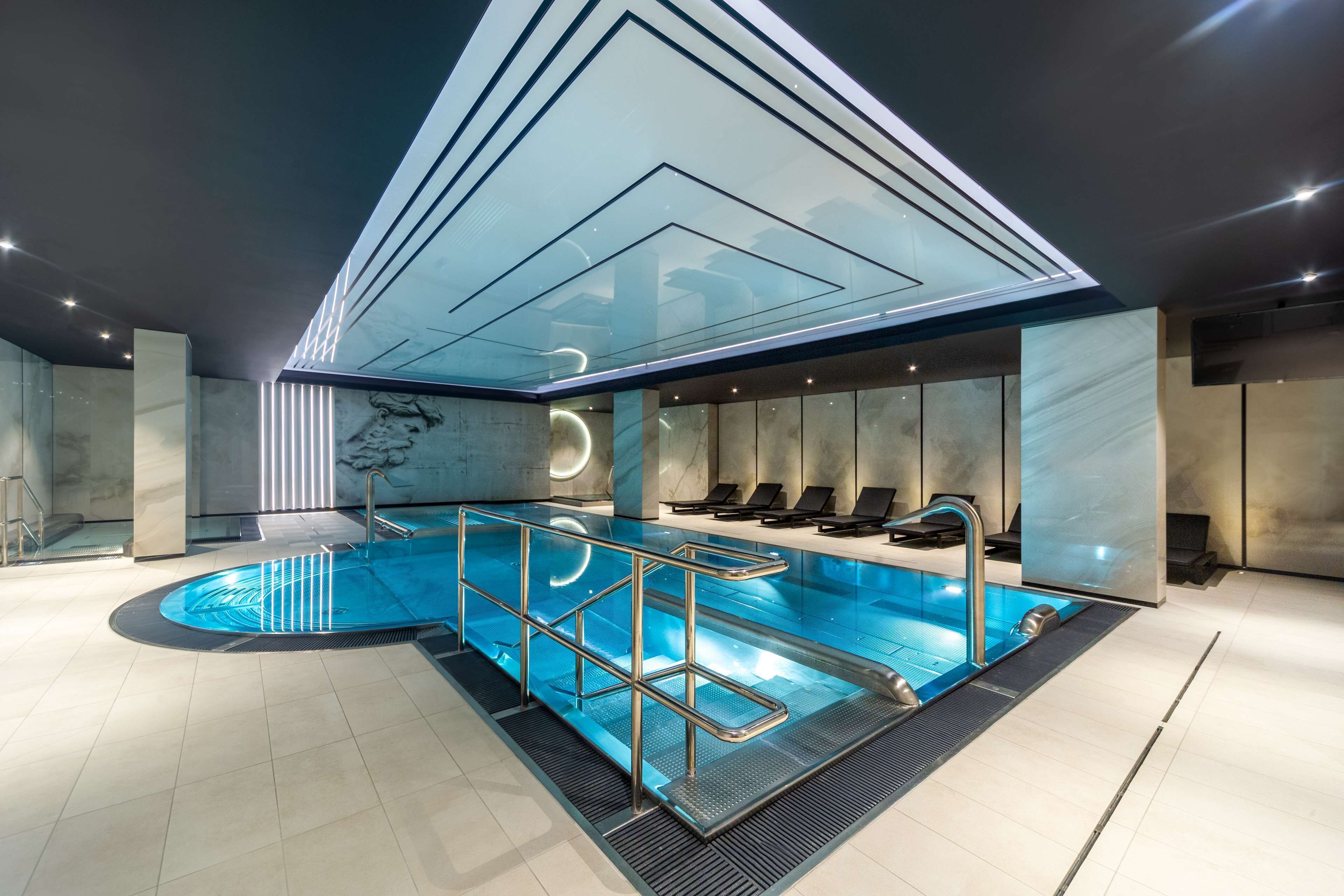 Ein moderner, überdachter Innenpool mit Glasdecke, Marmorwänden und Liegestühlen.