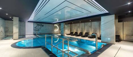 Piscina interior moderna con techo de cristal, paredes de mármol y sillones de descanso.