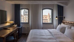 Ett hotellrum med en säng, ett skrivbord och två fönster med utsikt över byggnader.