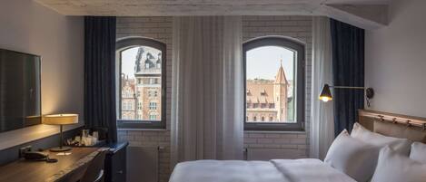Habitación de hotel con cama, escritorio y dos ventanas que dan vistas a edificios.