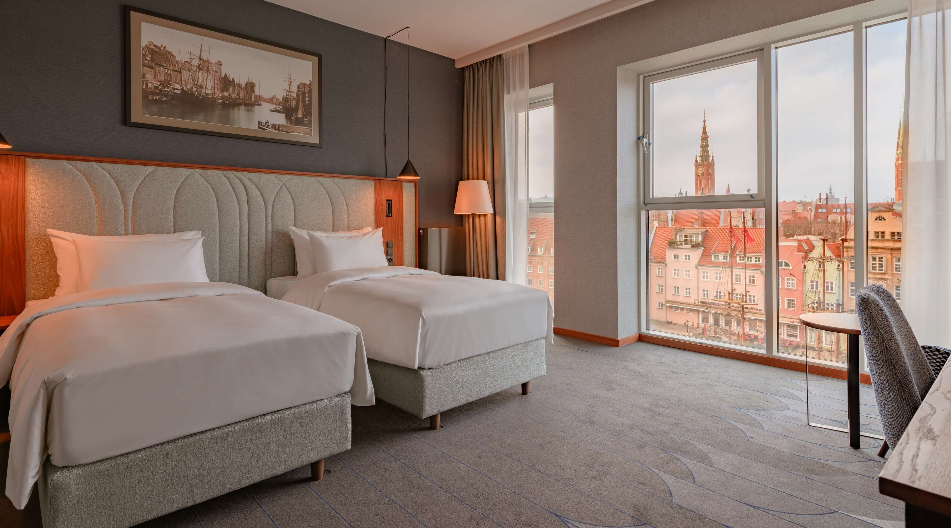 A hotel room with two beds, a large window with a city view, a framed picture on the wall, and a chair by the window.