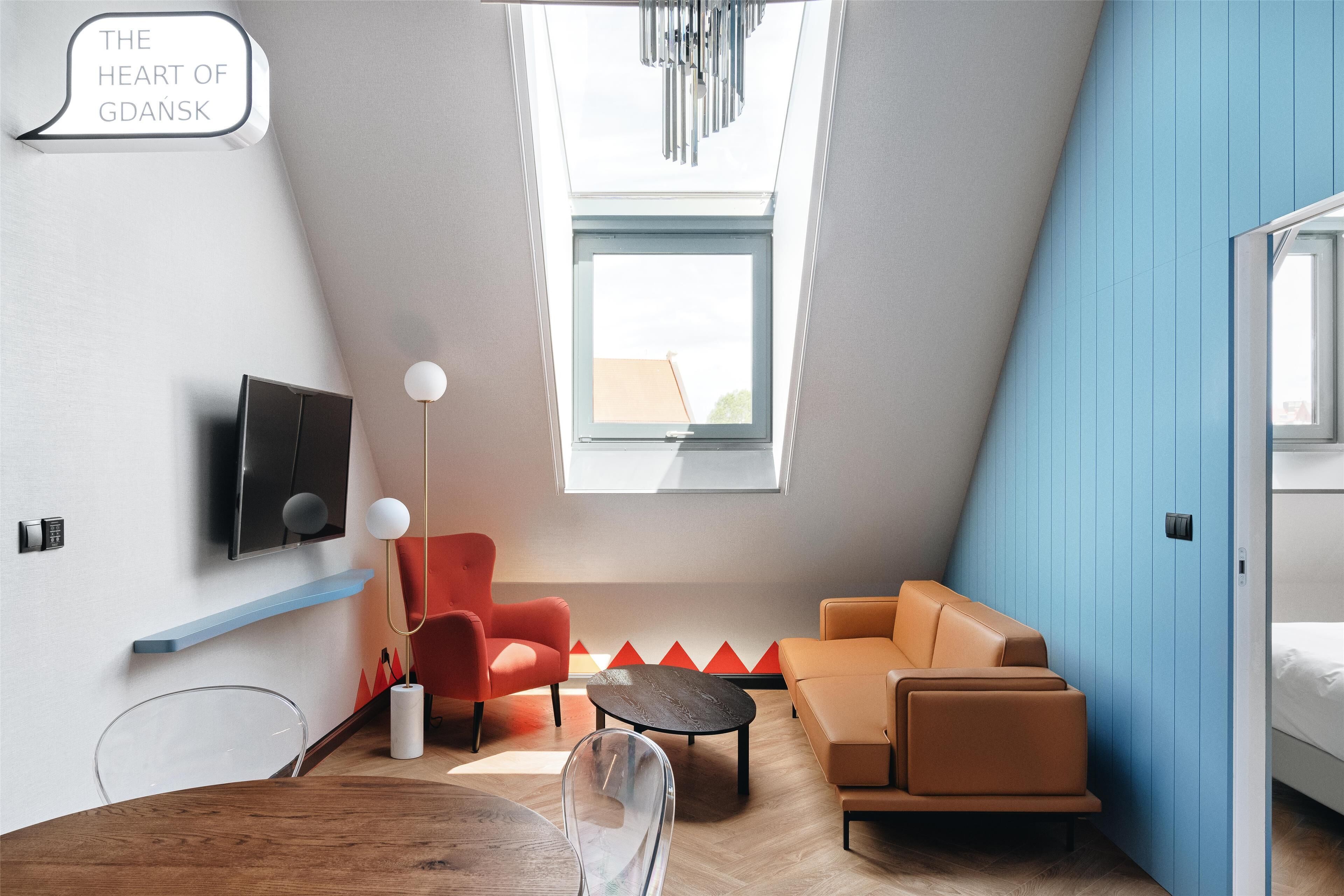 Une chambre moderne avec un plafond en pente, un téléviseur fixé au mur, un fauteuil rouge, un canapé orange et une petite table.