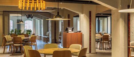 A spacious dining area with wooden tables and chairs, a central chandelier, and large windows allowing natural light.