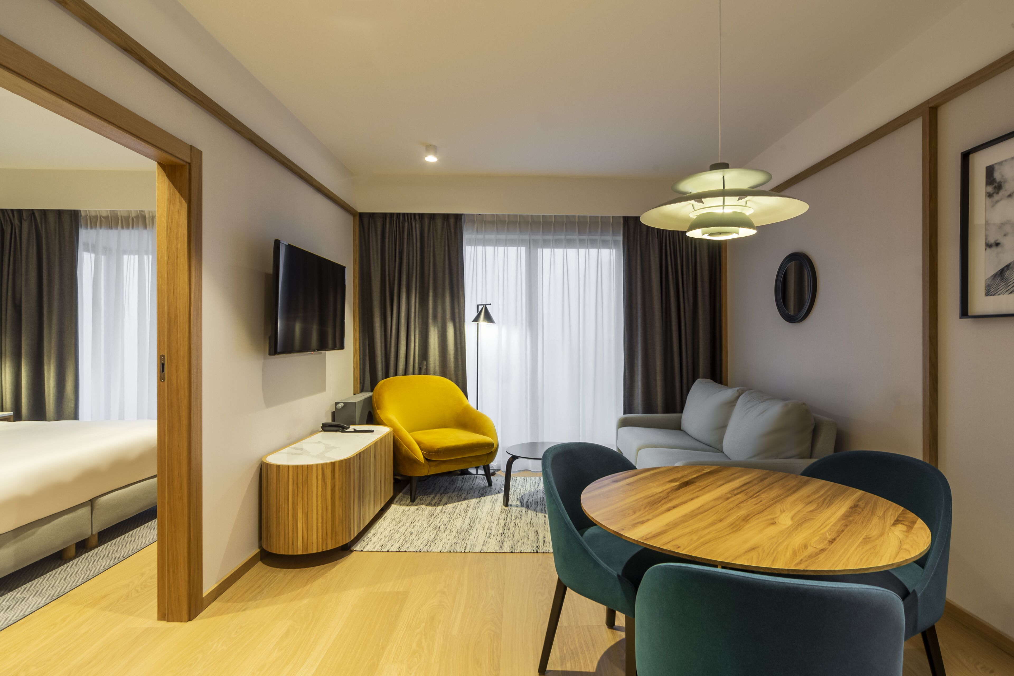 Ein modernes Hotelzimmer mit Holzboden, einem runden Holztisch, zwei blauen Stühlen, einem gelben Sessel, einem grauen Sofa, einem flachbildfernseher an der Wand und einem großen Fenster mit Vorhängen.