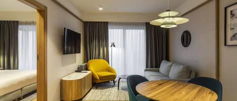 A modern hotel room with a wooden floor, a round wooden table, two blue chairs, a yellow armchair, a grey sofa, a flat-screen TV mounted on the wall, and a large window with curtains.