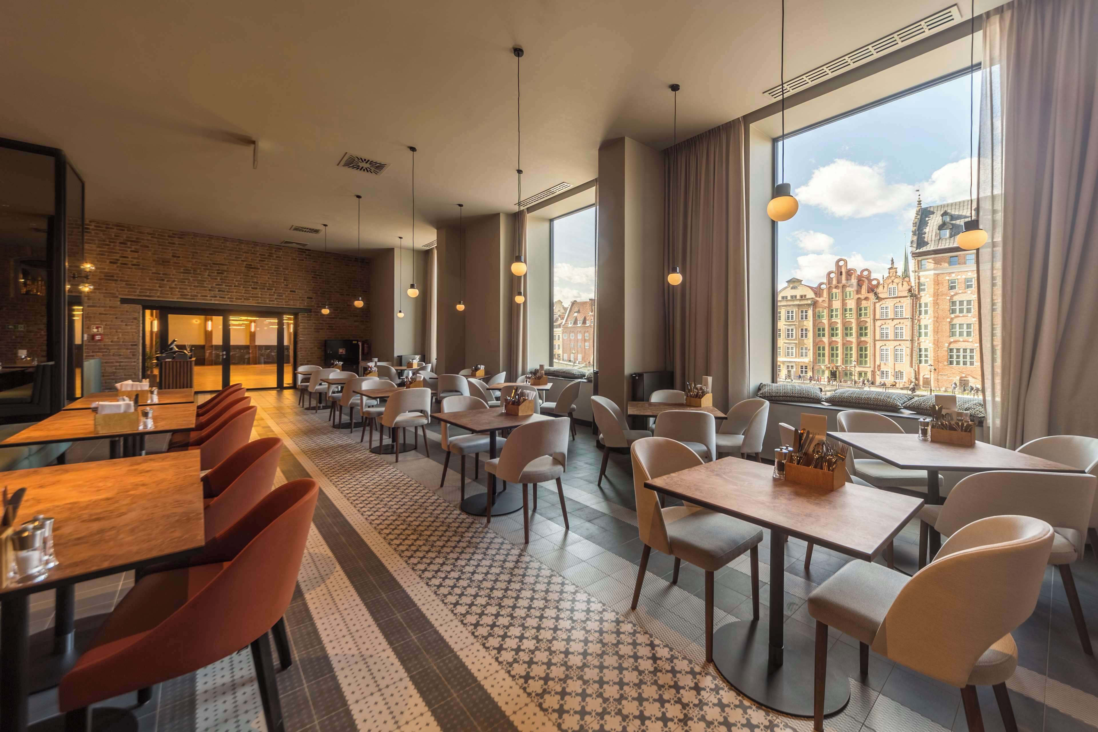A modern restaurant with high ceilings, exposed brick walls, and large windows offering city views. Tables are arranged in a spacious layout with ample seating.