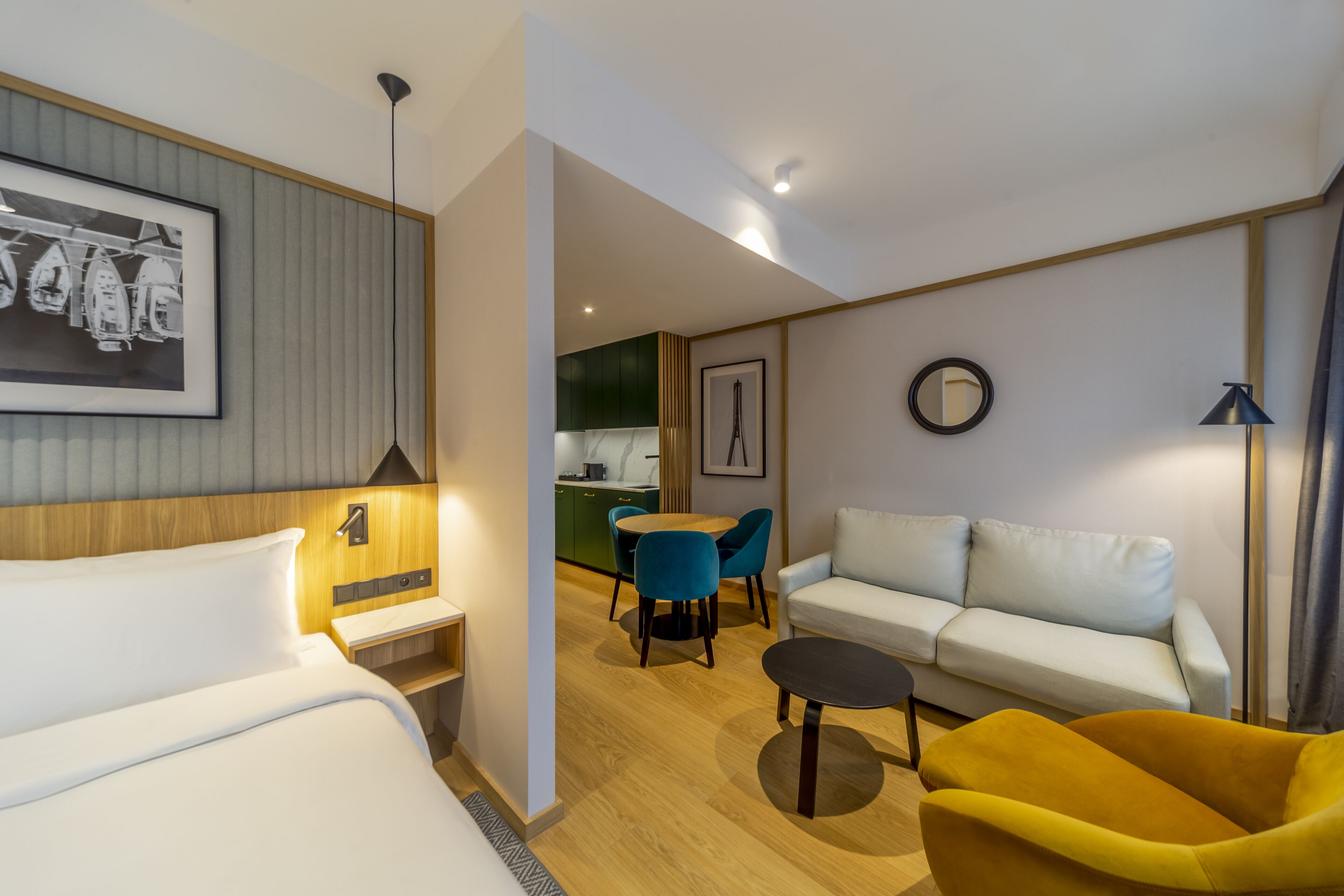 A modern hotel room with a bed, a sofa, a yellow armchair, a dining area with chairs, a kitchen in the background, and a framed picture on the wall.