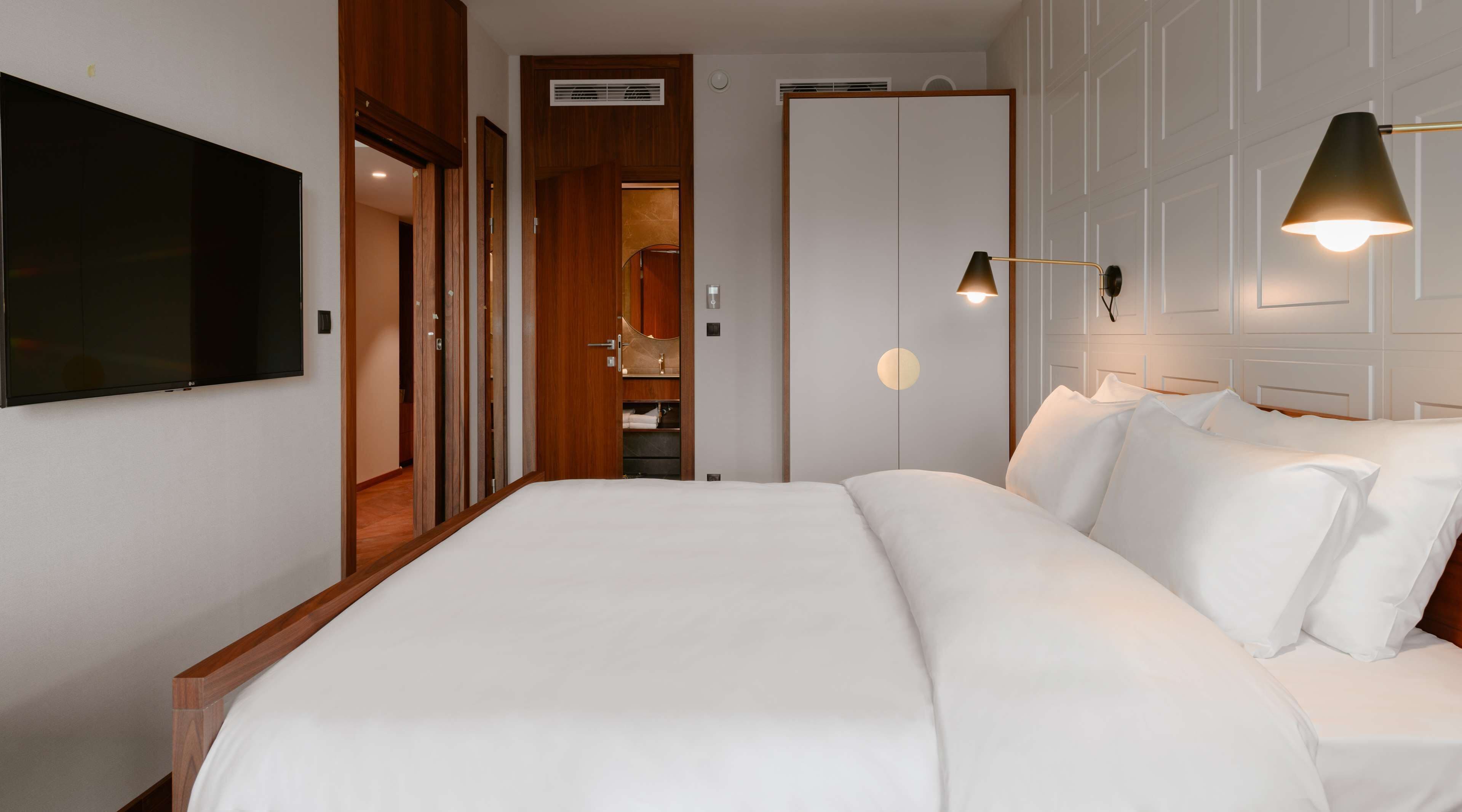 A hotel room with a large bed, a TV mounted on the wall, a wardrobe, and a bedside lamp.