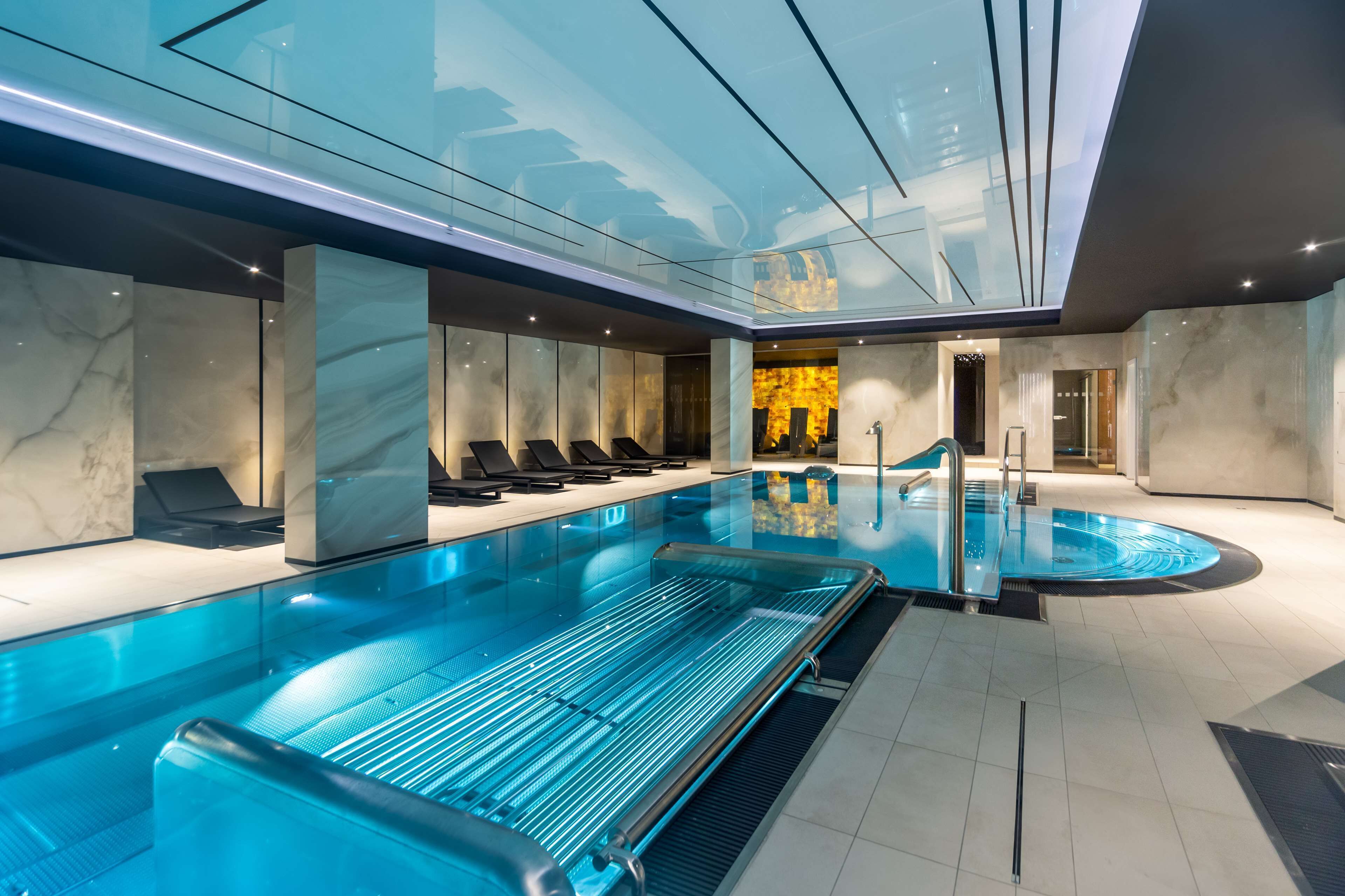 A modern indoor swimming pool with a glass ceiling, marble walls, and sun loungers.