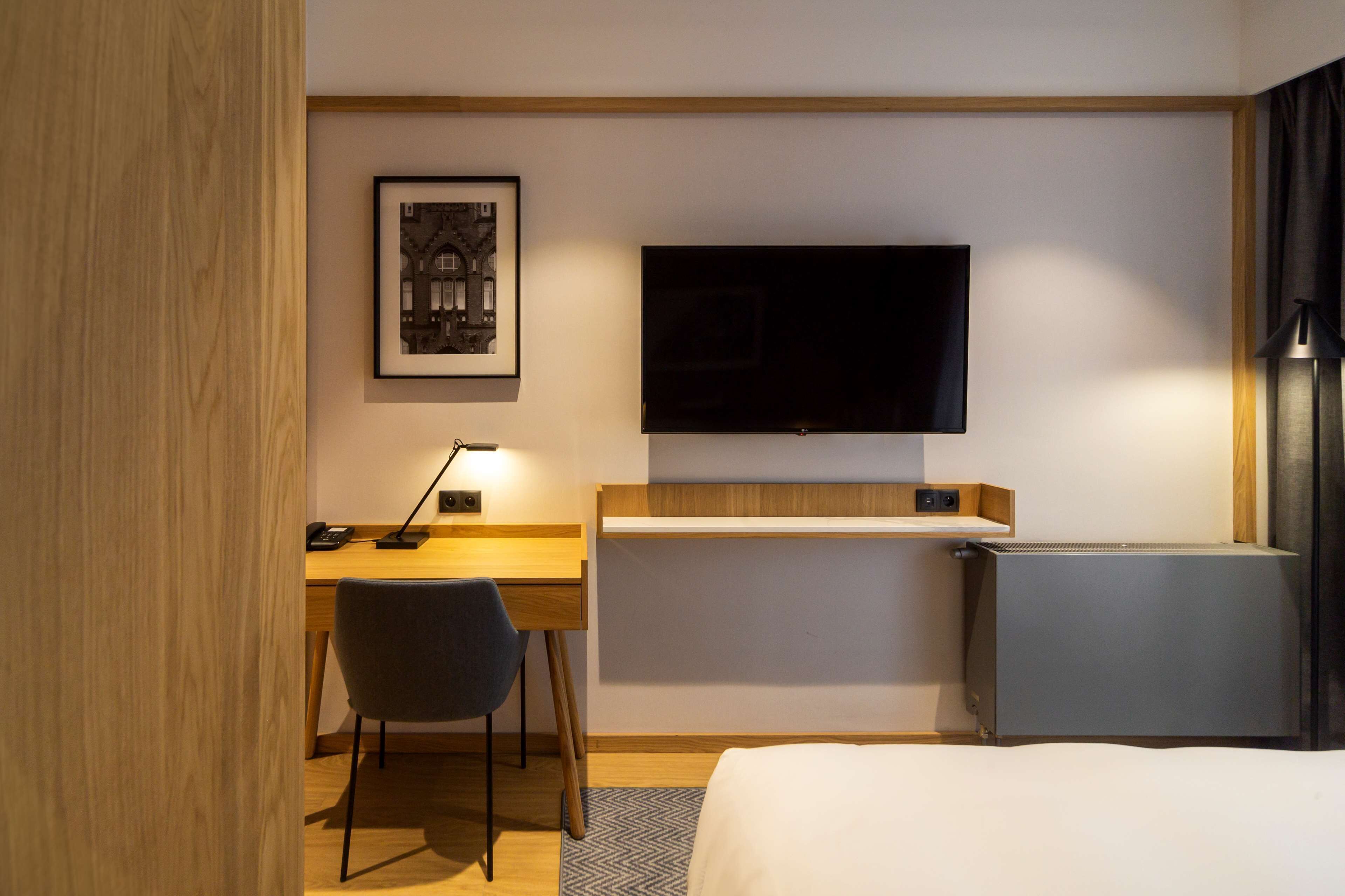 A hotel room with a bed, a desk with a chair, a flat-screen TV mounted on the wall, and a small refrigerator.