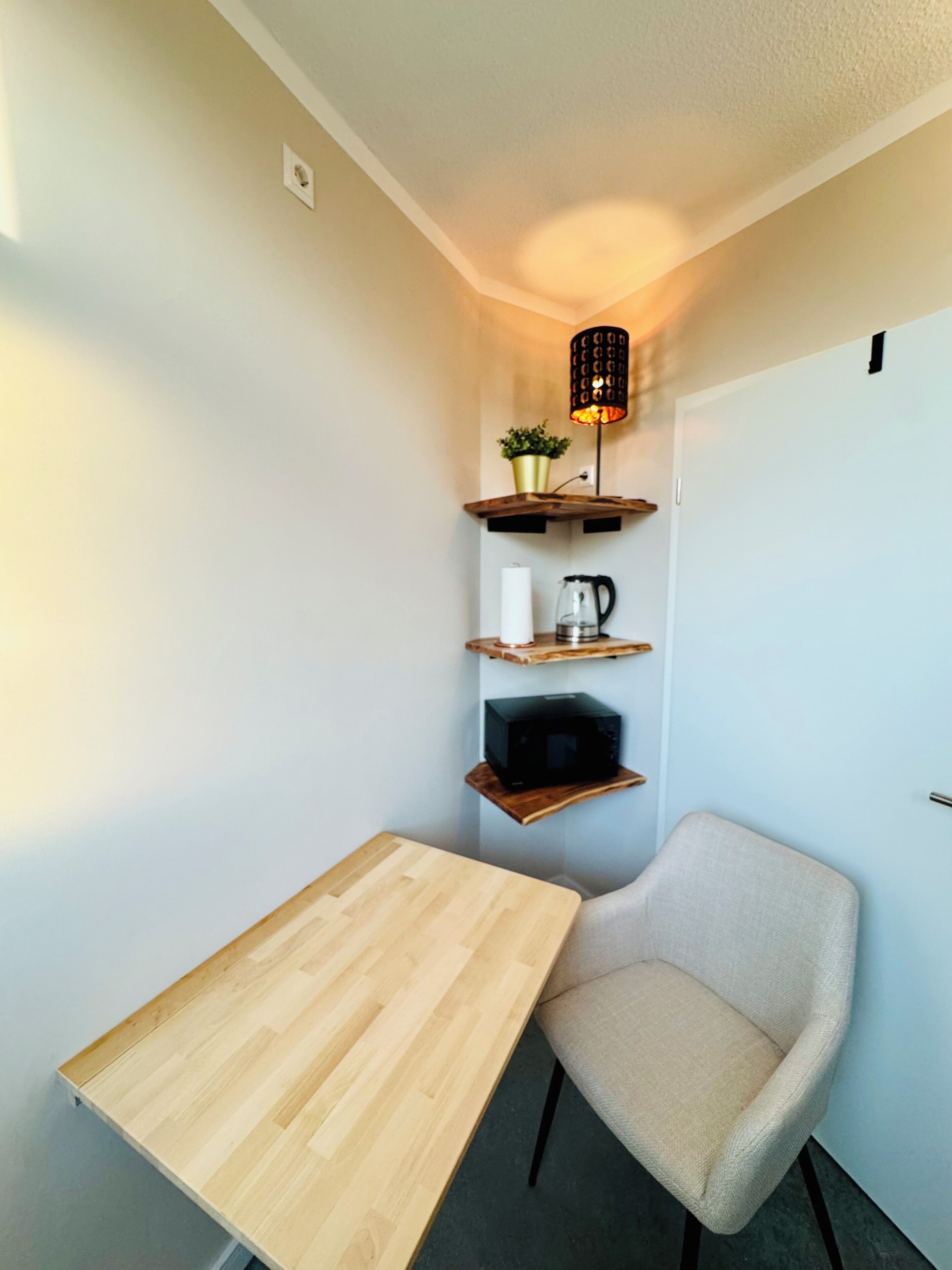 A small room with a wooden table, a chair, and shelves with a lamp, a plant, and a kettle.