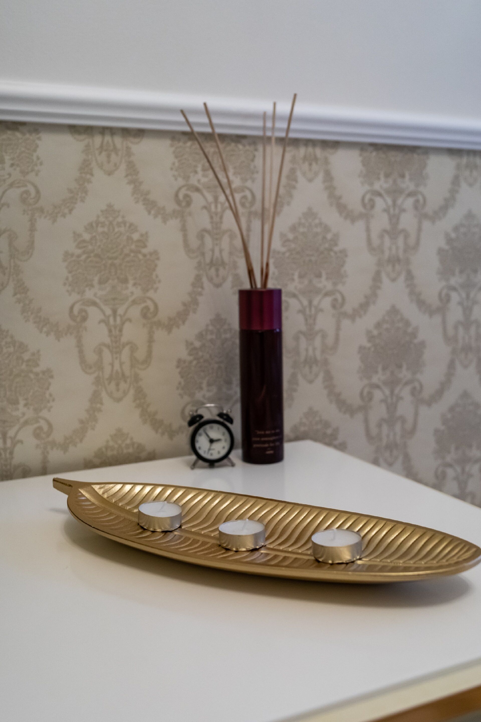 A decorative tray with three candles on a white table against a patterned wallpaper background.