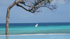 A clear view of a beach with a sailboat on the water, a tree in the foreground, and a clear sky. - Karamba (Kizimkazi)