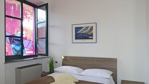 A bedroom with a bed, a nightstand, a bedside table, a window with a view of trees and mountains, and a framed picture of a sailboat on the wall. - Holideal Campione Beach House (Tremosine)