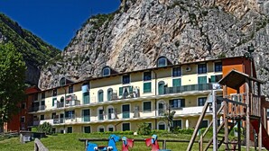 A playground with swings and slides, a wooden fence, and a building with balconies in the background. - Holideal Campione Beach House (Tremosine)