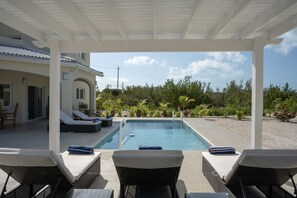 Área de piscina coberta ao ar livre com espreguiçadeiras e vista para uma casa e vegetação.