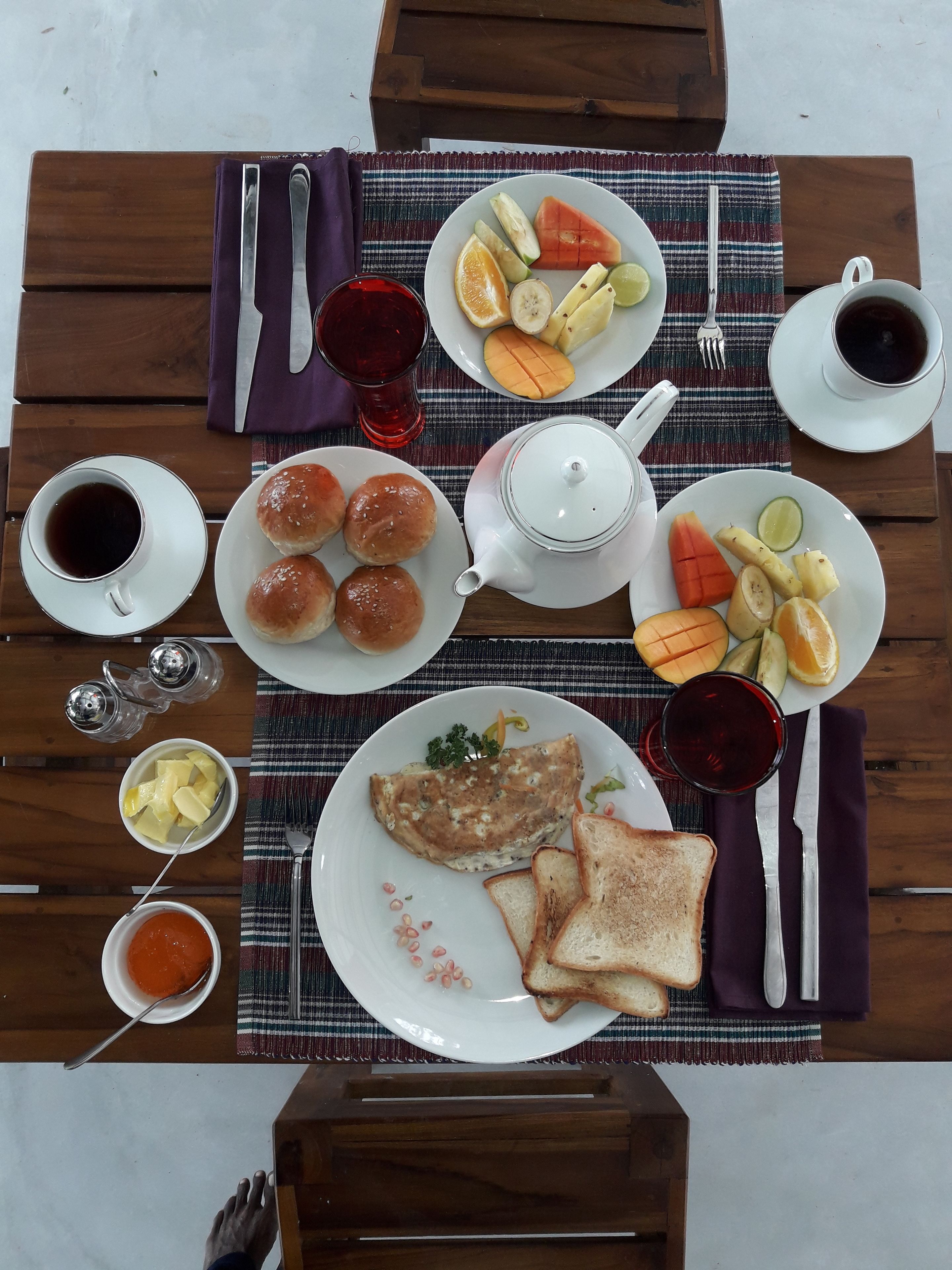 Une table dressée pour le petit-déjeuner, avec une variété d’aliments, notamment des toasts, des œufs et des fruits, disposés sur une table en bois surmontée d’une nappe à rayures.