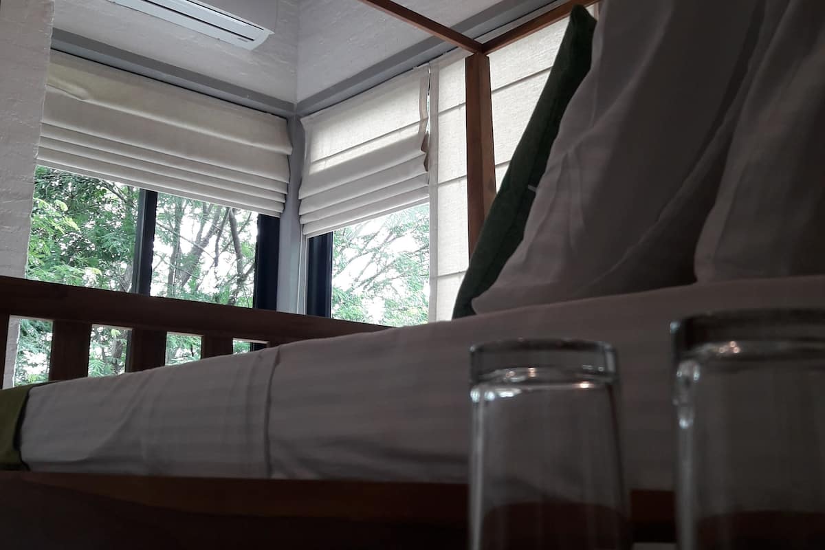 Una cama con sábanas blancas, una mesita de noche con vasos y una ventana con persianas.