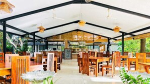 Un espace de restauration intérieur avec des tables et des chaises en bois, un plafond haut agrémenté de luminaires suspendus et une vue sur l’extérieur.