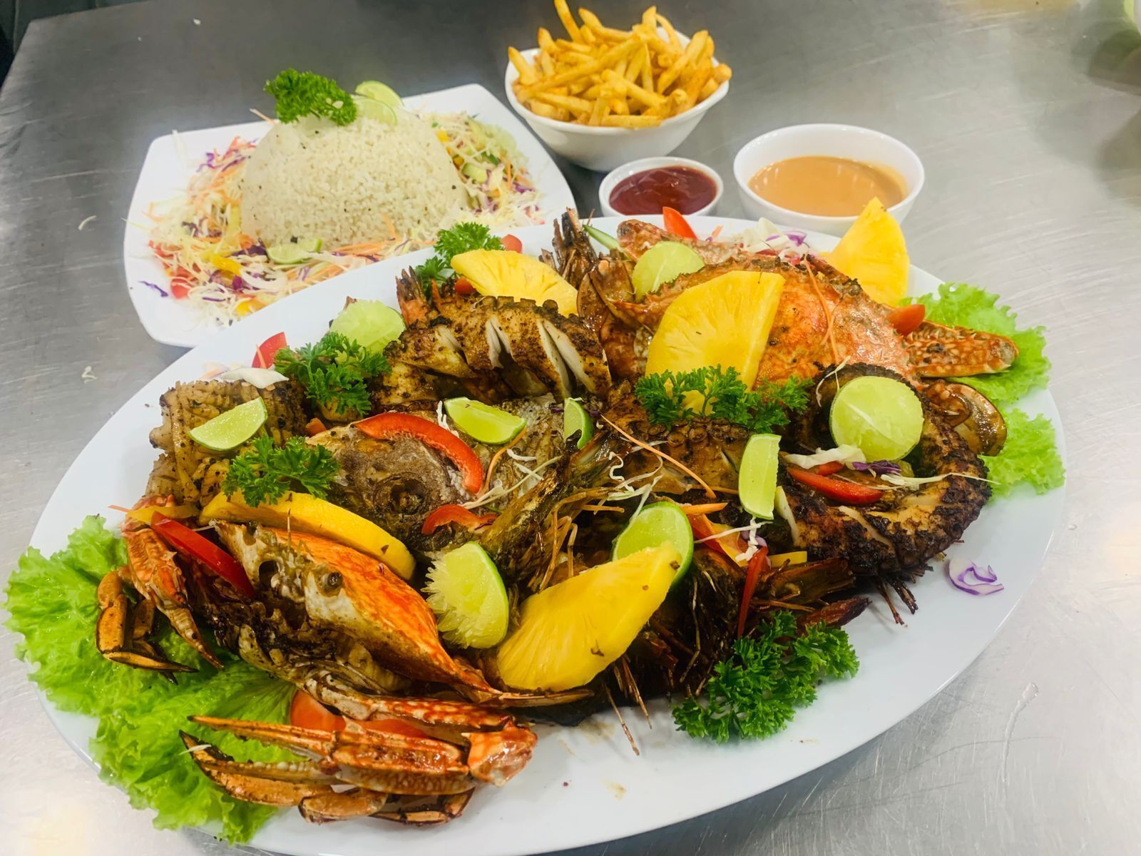 Una bandeja de mariscos con pescado a la parrilla, cangrejo y verduras, acompañada de arroz, papas fritas y salsas para mojar.