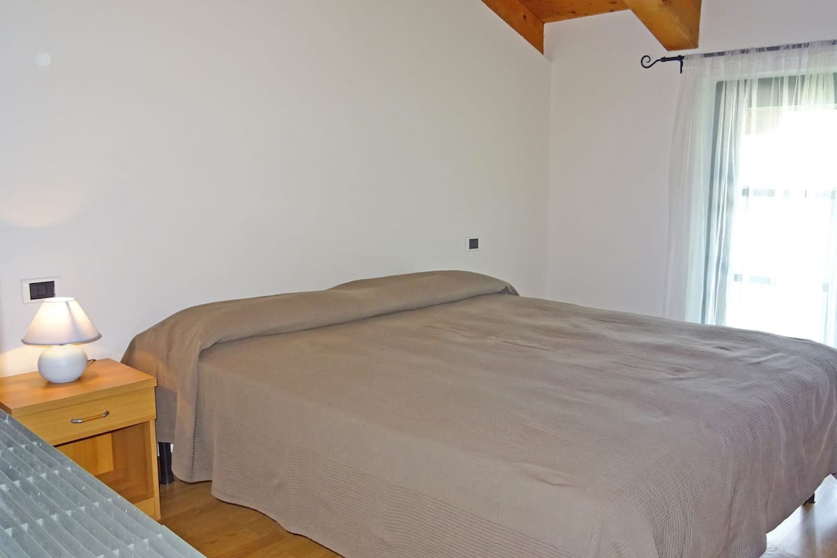 Ein Schlafzimmer mit einem Bett, einem Nachttisch mit Lampe, einem Fenster mit Vorhängen und einer Holzbalkendecke.