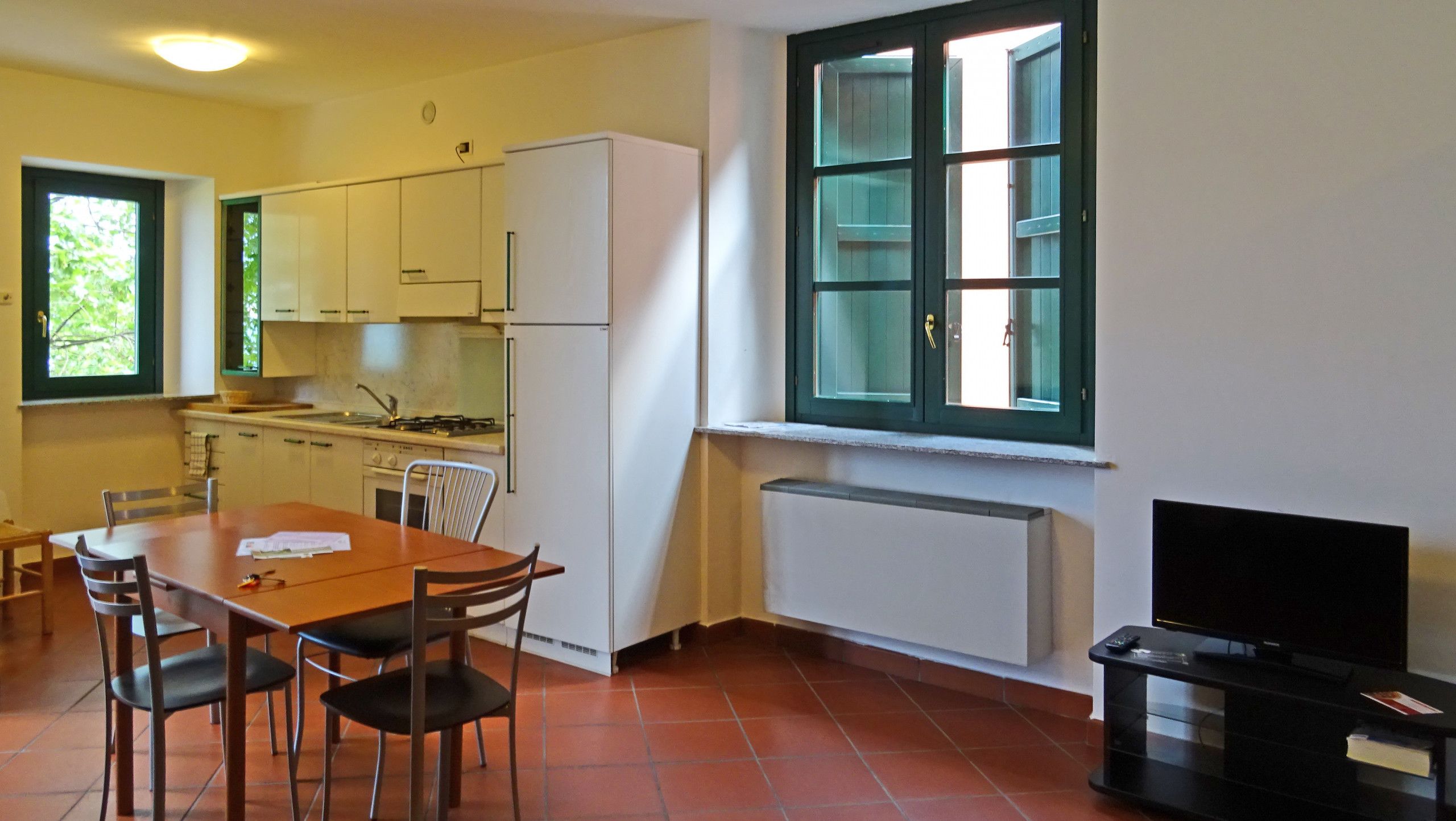 A compact kitchen with white cabinets, a dining table with chairs, a built-in stove, and a window with green shutters.