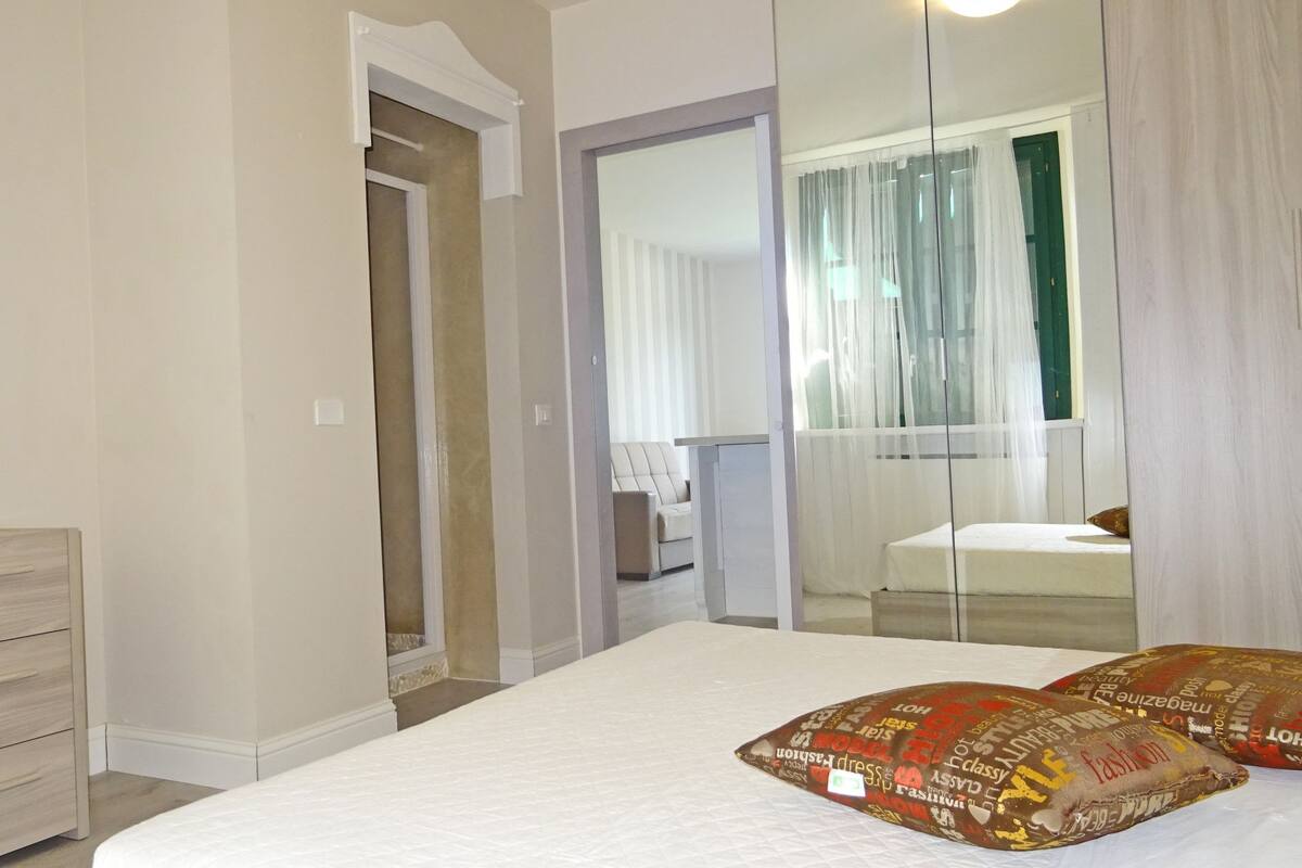 Quarto com uma cama grande, cômoda, espelho e vista para uma varanda com porta verde.