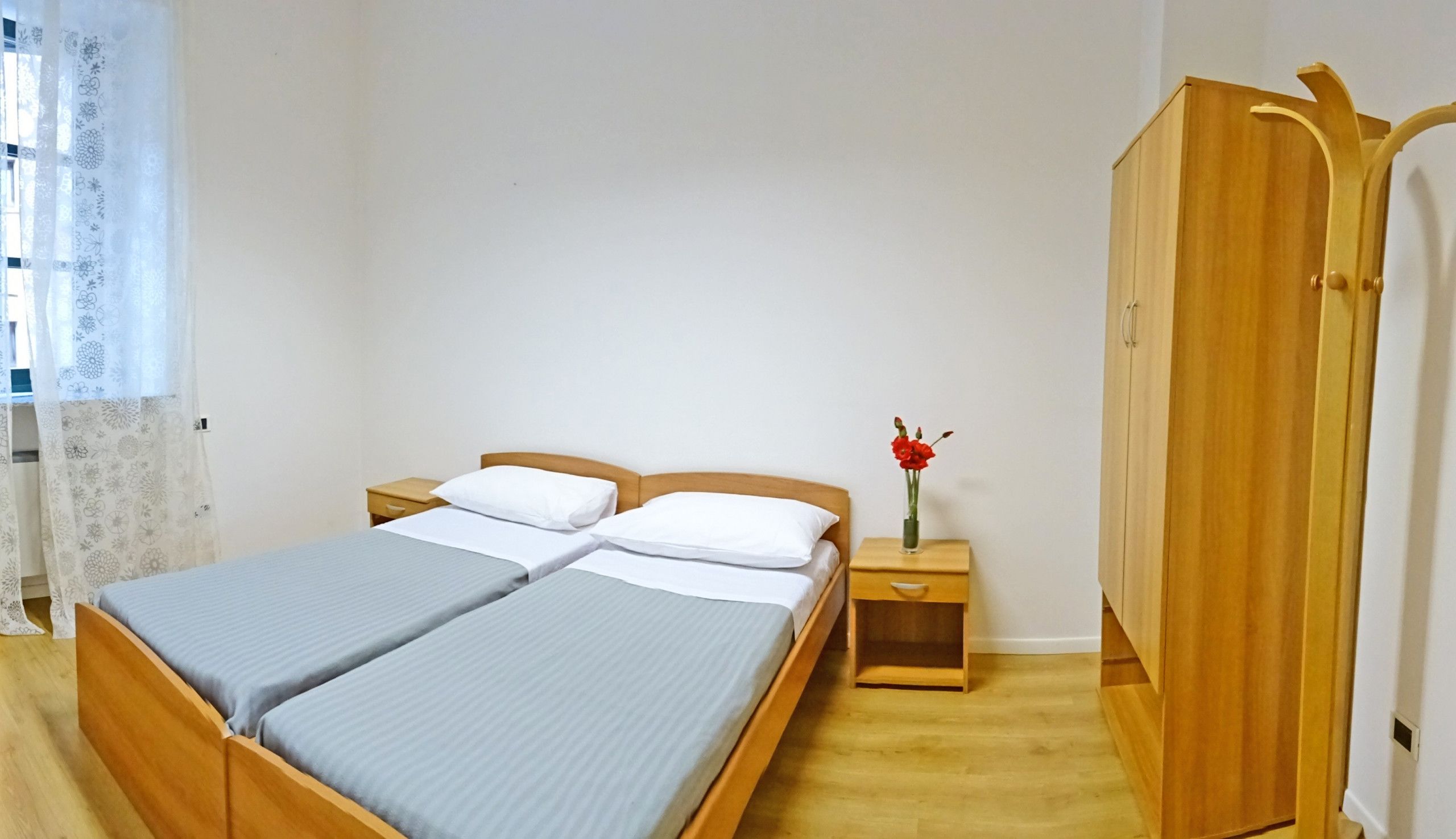 Een tweepersoonsbed met houten frame, twee nachtkastjes, een houten garderobe en een raam met dunne gordijnen.