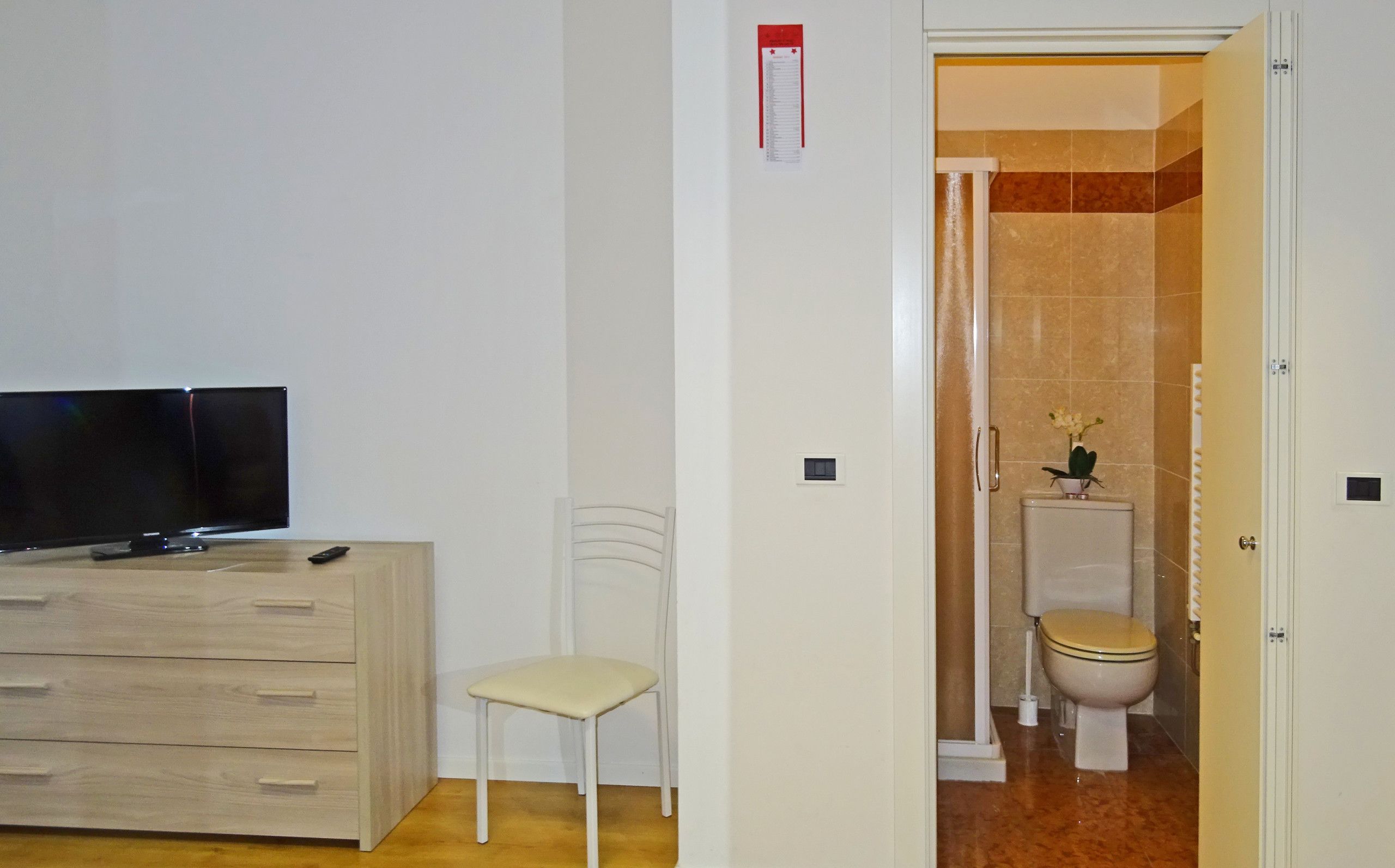 Ein Zimmer mit Fernseher auf einer Holzkommode, einem weißen Stuhl und einem Badezimmer mit Toilette und Dusche.