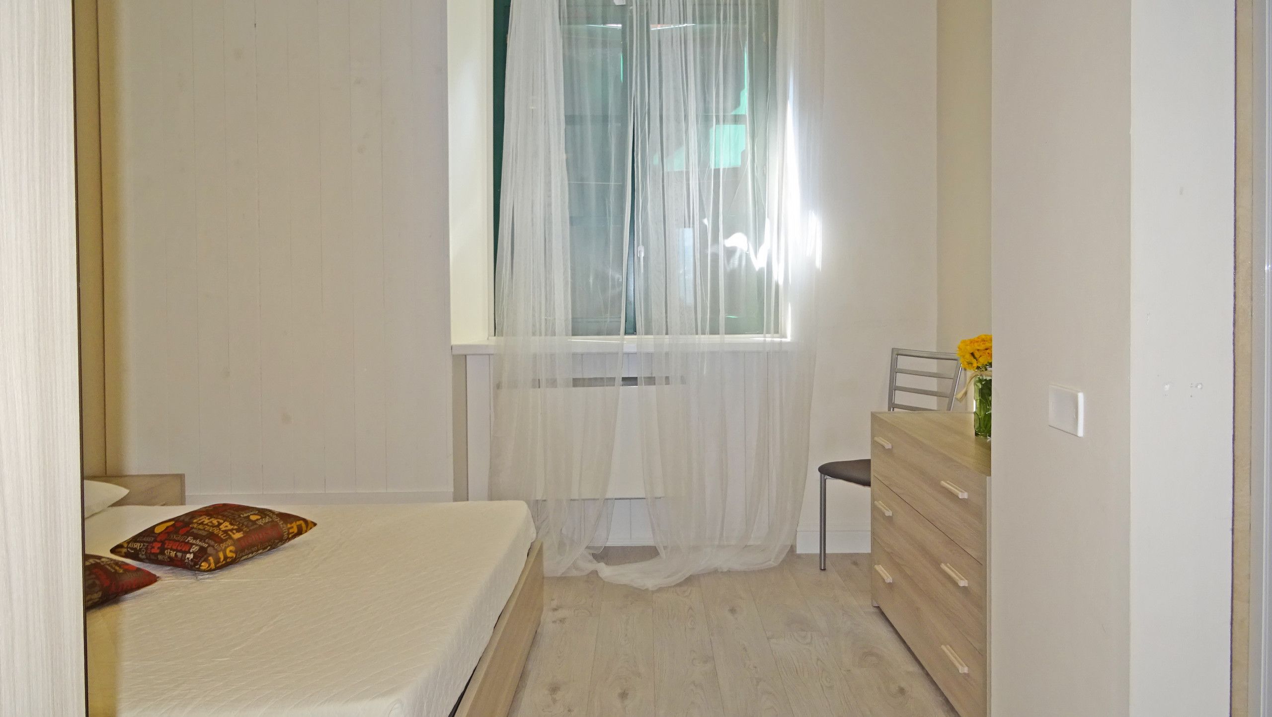 Un dormitorio con cama, una silla, cómoda y ventana con cortinas traslúcidas.
