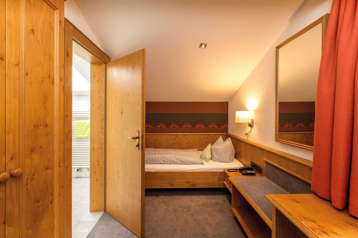 Una habitación con una cama de madera, una mesita de noche, una lámpara, un espejo y una puerta que da a otra estancia.