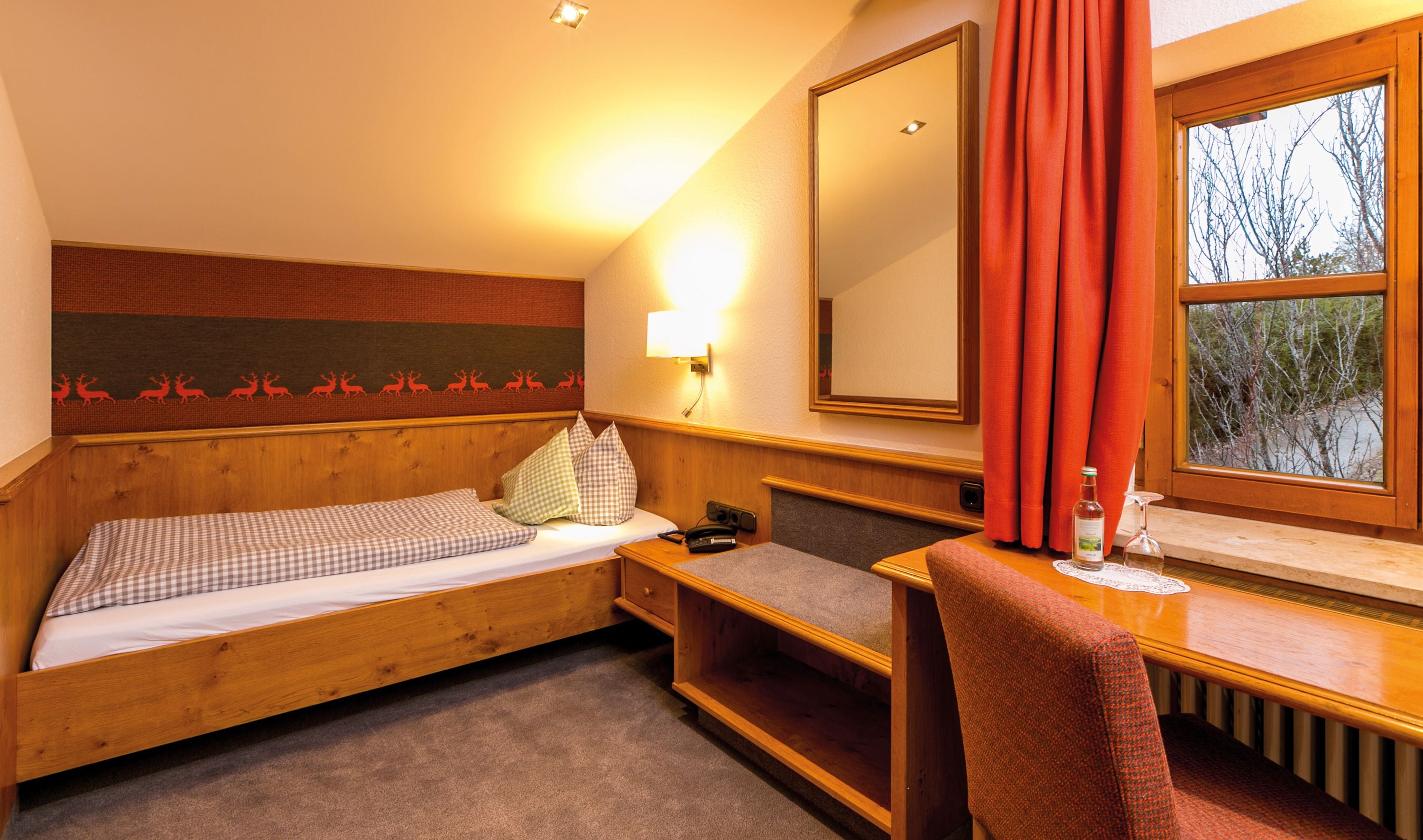 A single bed with a checkered bedspread, a bedside table with a lamp, a chair, a small desk with a bottle, and a window with curtains.