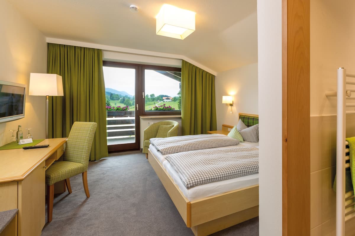 Habitación de hotel con escritorio, dos sillas, cama con ropa de cama a cuadros, ventana con cortinas y vista a zonas verdes.