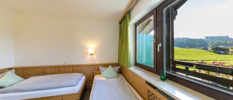 A room with two wooden beds, a window with a view of a green landscape, and a wall-mounted light.