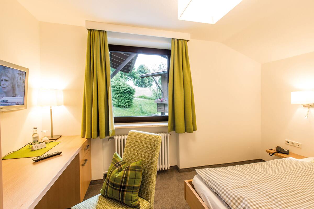 Habitación de hotel con una cama, una silla con almohada, un escritorio con lámpara y una ventana con cortinas.