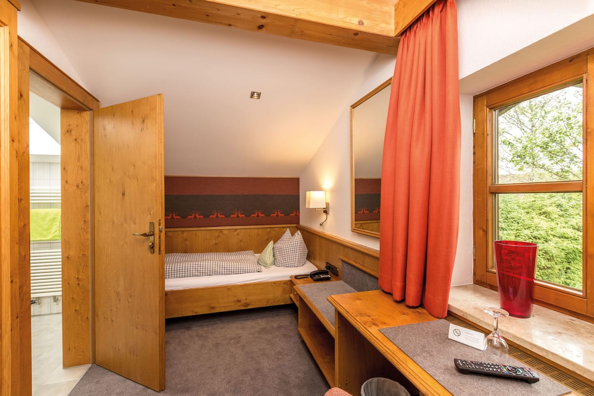 Una habitación con estructura de madera, que incluye una cama, un banco, una mesa, un vaso de vidrio rojo y una ventana con vistas a la vegetación.
