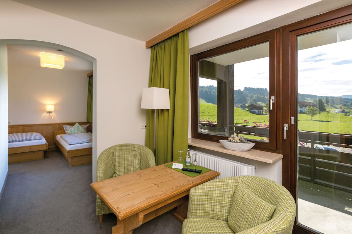 Una habitación con una mesa de madera, dos sillas verdes a cuadros, un sofá y una ventana que da a un paisaje verde.