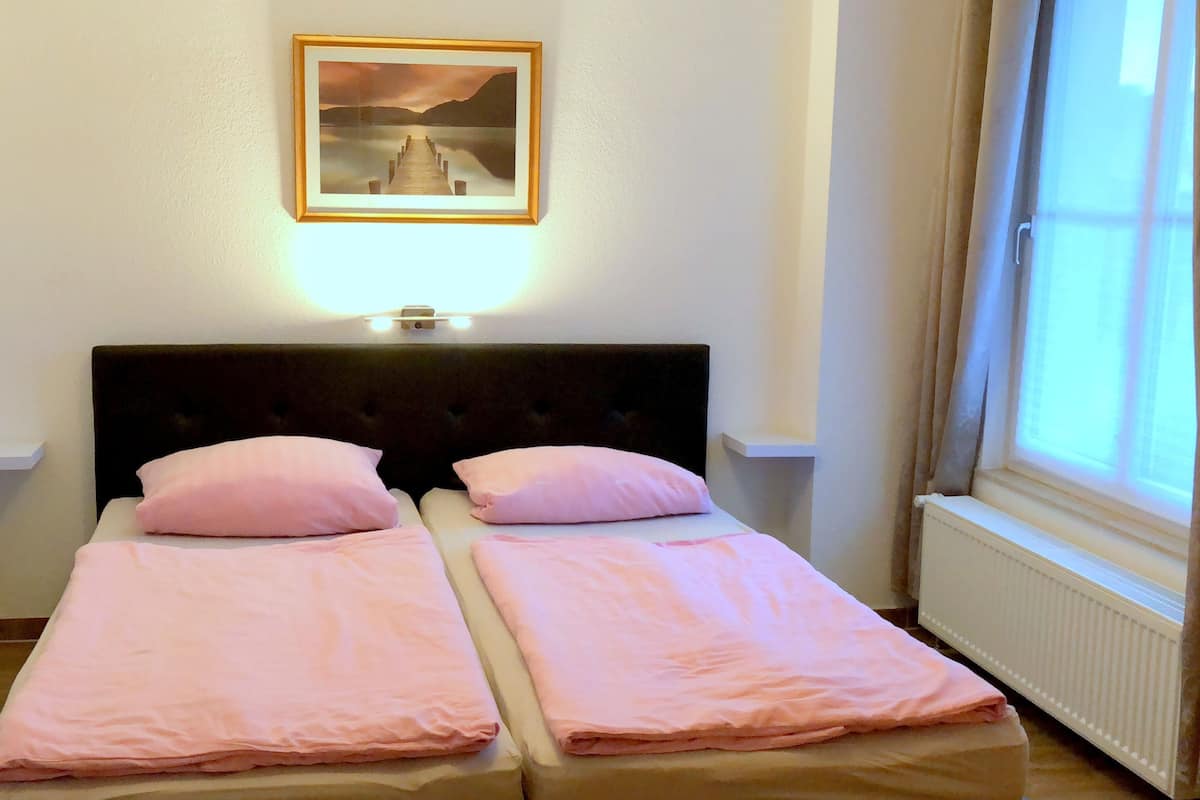 A double bed with two pink pillows and a headboard, a framed picture above the bed, and a window with curtains on the right.