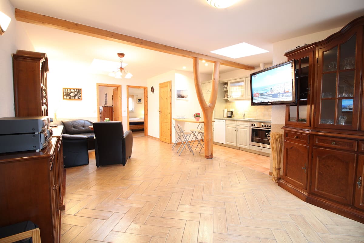 A spacious living room with a wooden herringbone floor, a television mounted on a wooden cabinet, and a dining area with a table and chairs.
