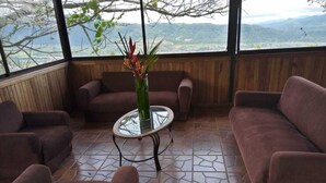 A seating area with a glass coffee table, cushioned sofas, and a view of a mountainous landscape through large windows. - Hotel Green Mountain Turrialba (Turrialba)