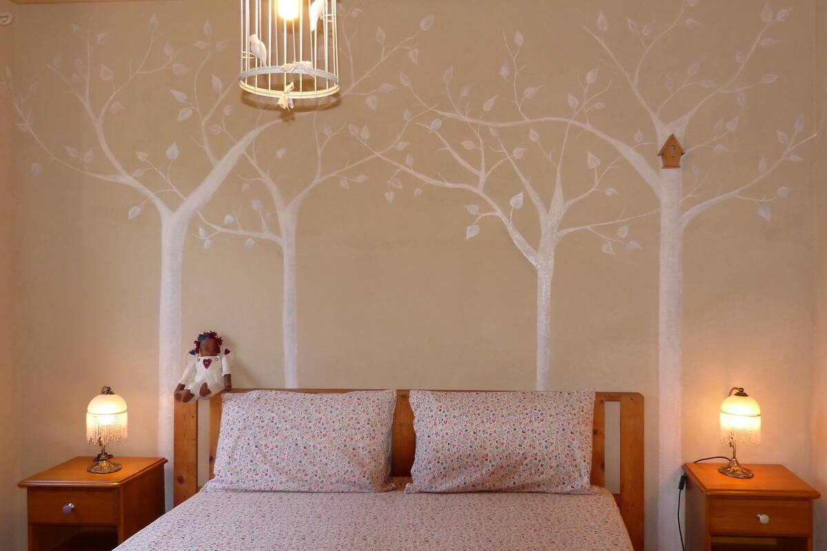 A bedroom with a bed, two nightstands, a hanging light fixture, and a wall mural of trees.