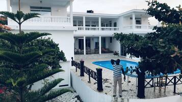 A modern white building with a swimming pool, surrounded by lush greenery and a stone pathway.