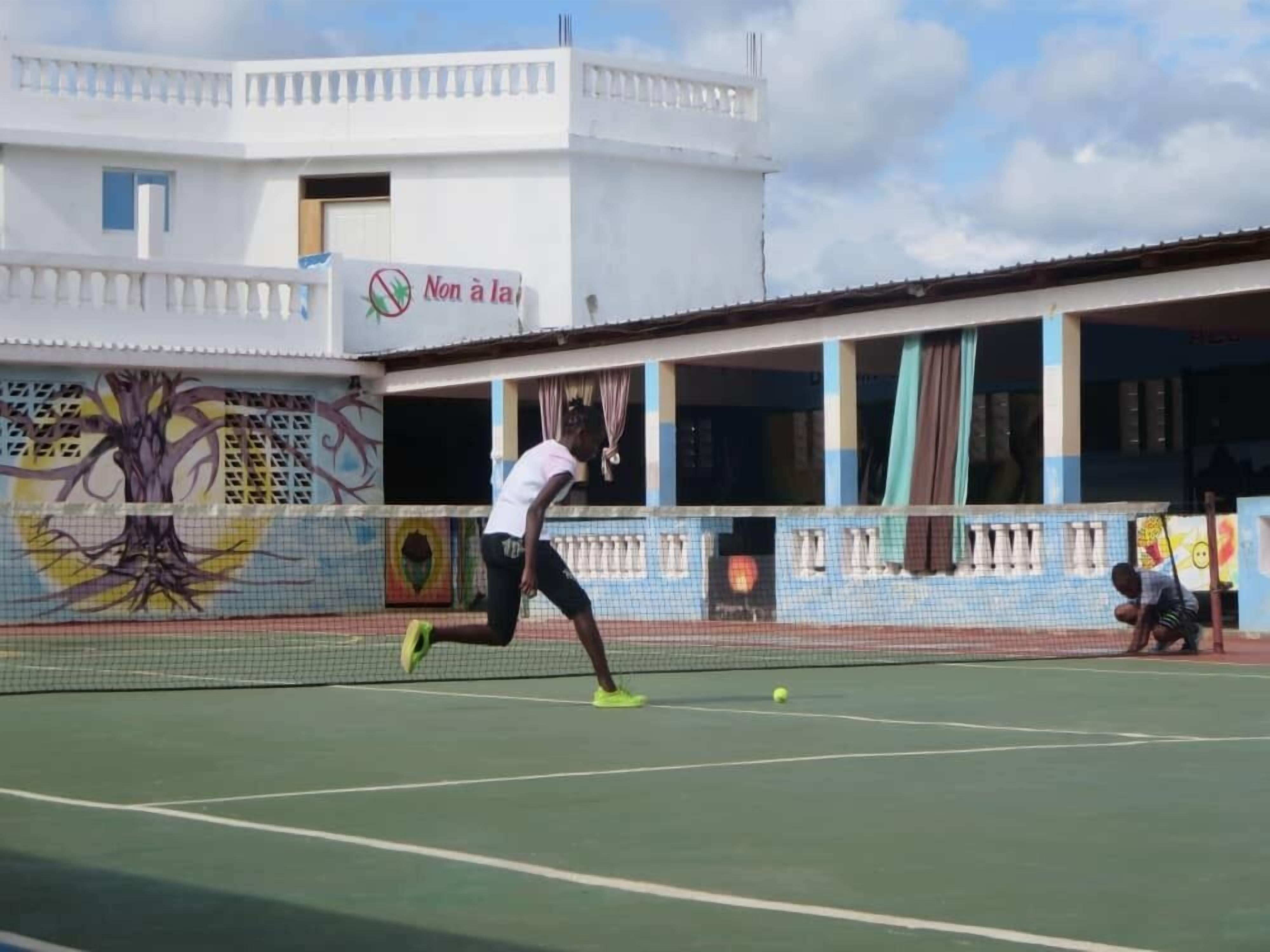 A tennis court with a net, a player in motion, and a building with a mural in the background.