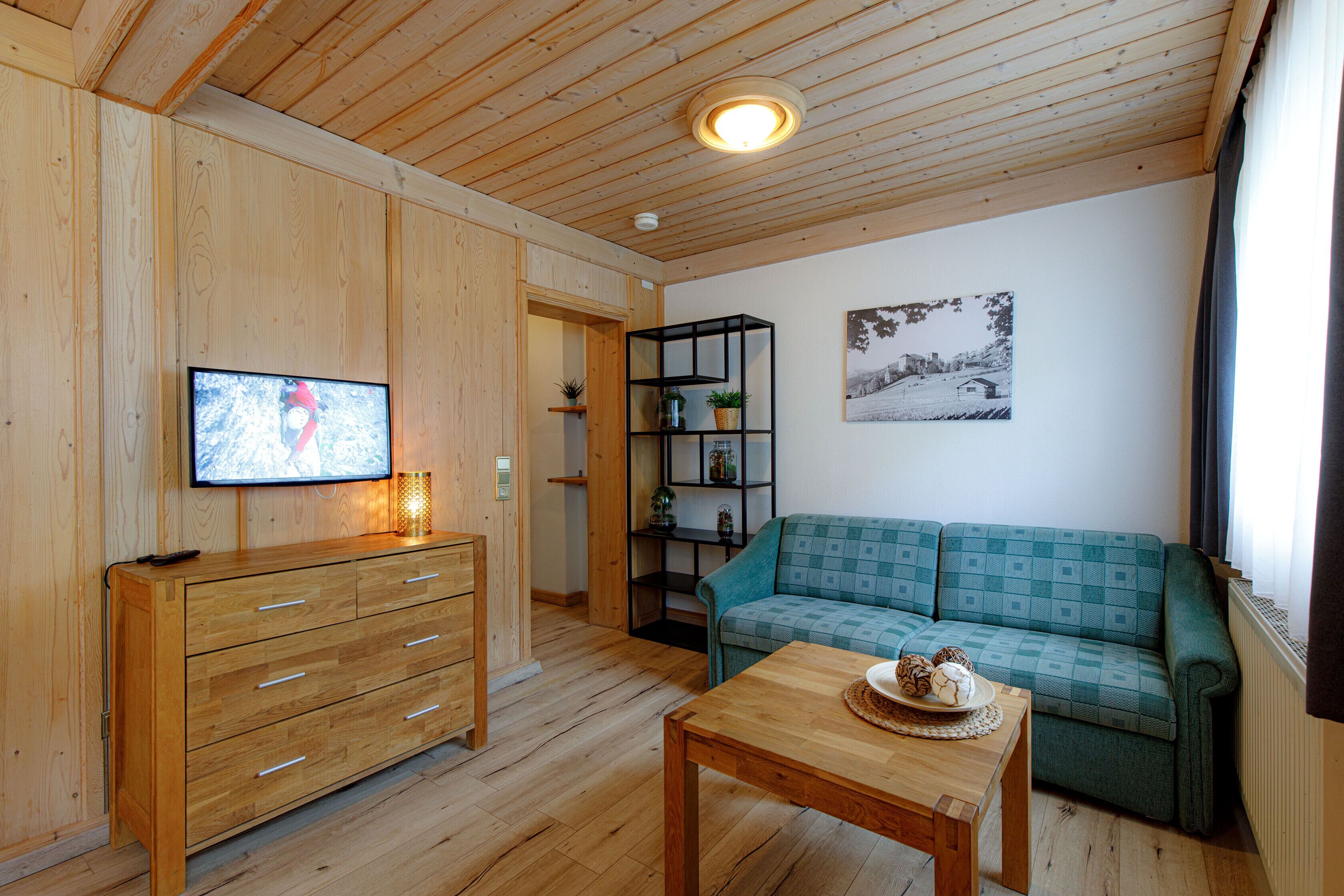 Une pièce aux murs lambrissés, avec une commode en bois, un canapé vert, une table en bois sur laquelle se trouve une assiette de biscuits, une étagère noire et un tableau encadré accroché au mur.